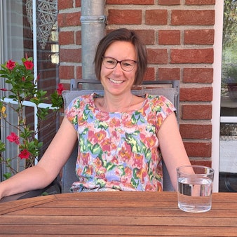 Eine Frau sitzt auf ihrer Terrasse an einem Tisch mit einem Glas Wasser.