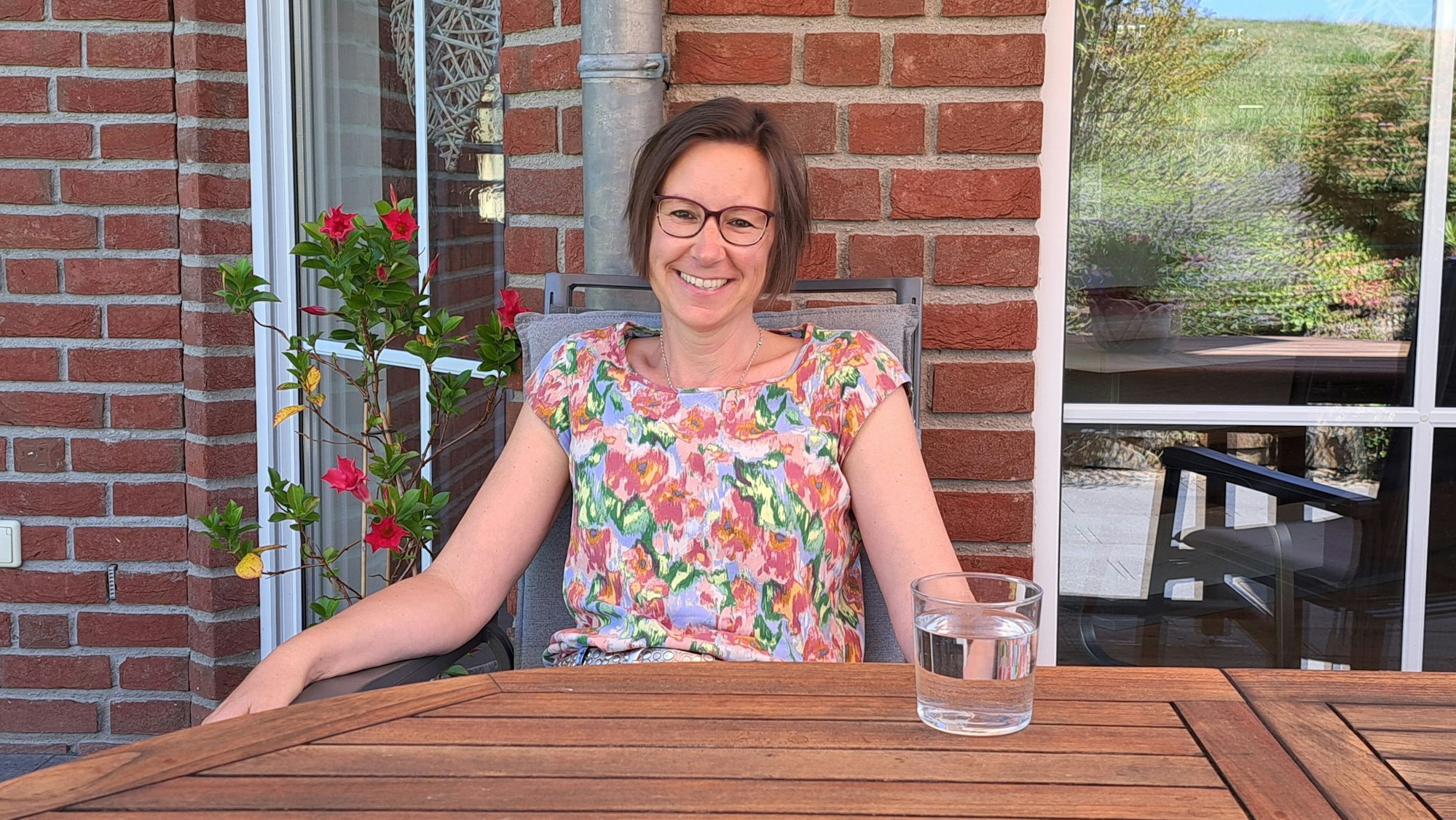 Eine Frau sitzt auf ihrer Terrasse an einem Tisch mit einem Glas Wasser.