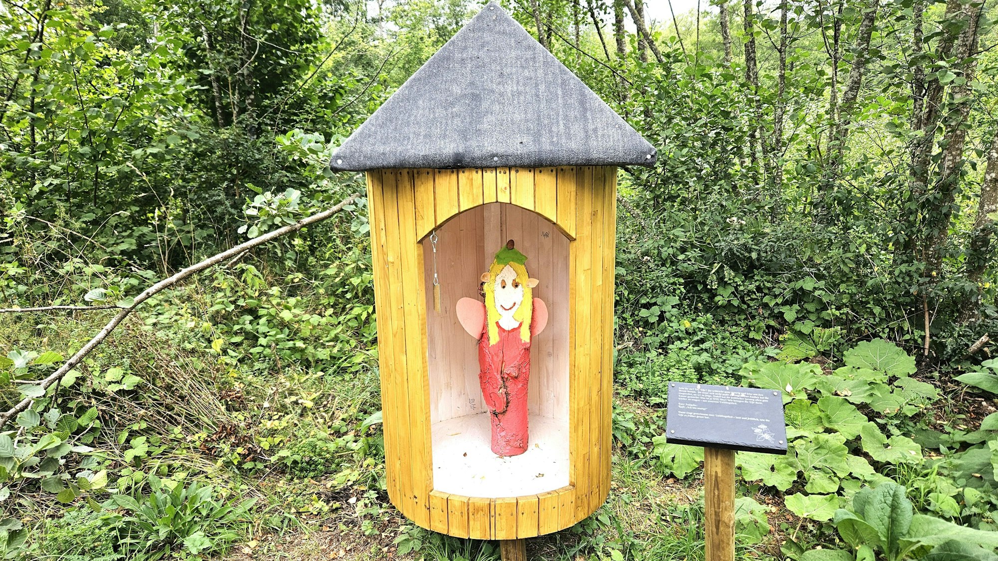 Eine Elfenfigur mit rotem Kleid steht in einer runden Holzhütte mit spitzem Dach.