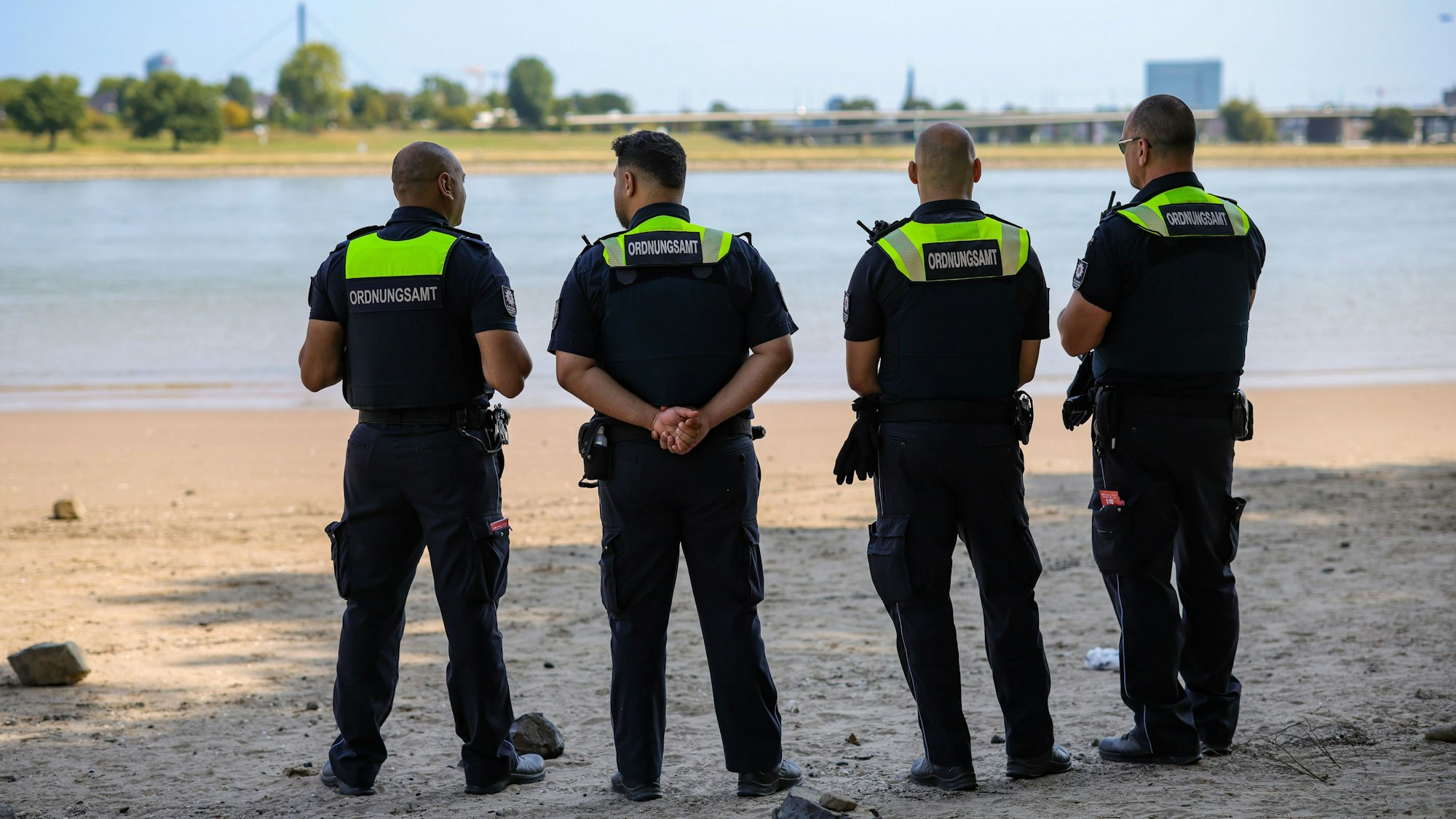 Vier Mitarbeiter vom Ordnungsamt der Stadt Düsseldorf stehen an einem Strandabschnitt des Rheins und überwachen das Badeverbot, das Düsseldorf am 14. August erlassen hat.