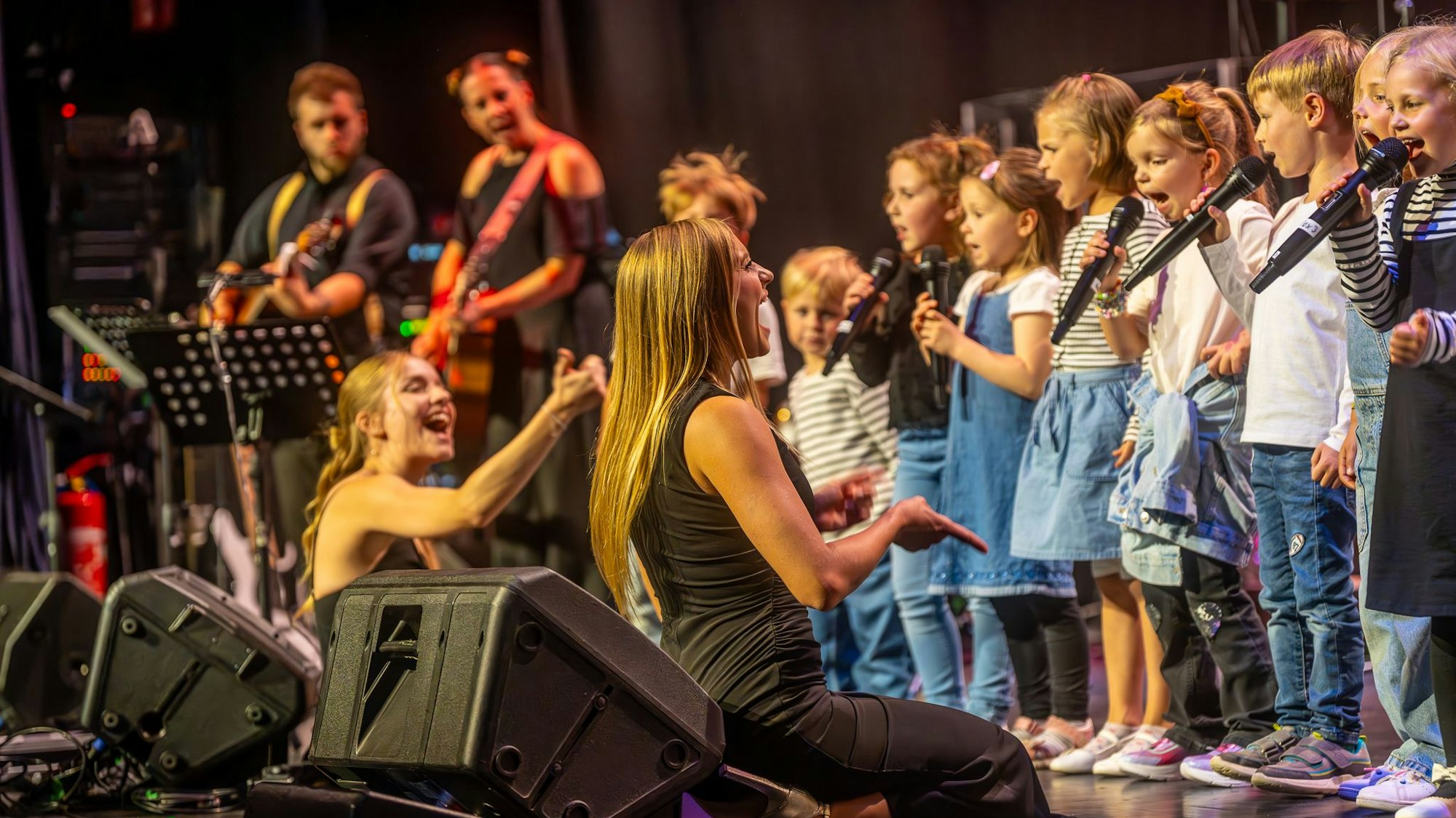 Das Bild zeigt einen Kinderchor, vor ihnen die Chorleitung auf den Knien, um den Chor zu animieren.