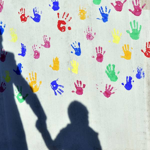 Ein Mann mit einem Kind auf dem Arm und einem an der Hand wirft einen Schatten auf eine mit bunten Handabdrücken bemalte Wand einer Kindertagesstätte.