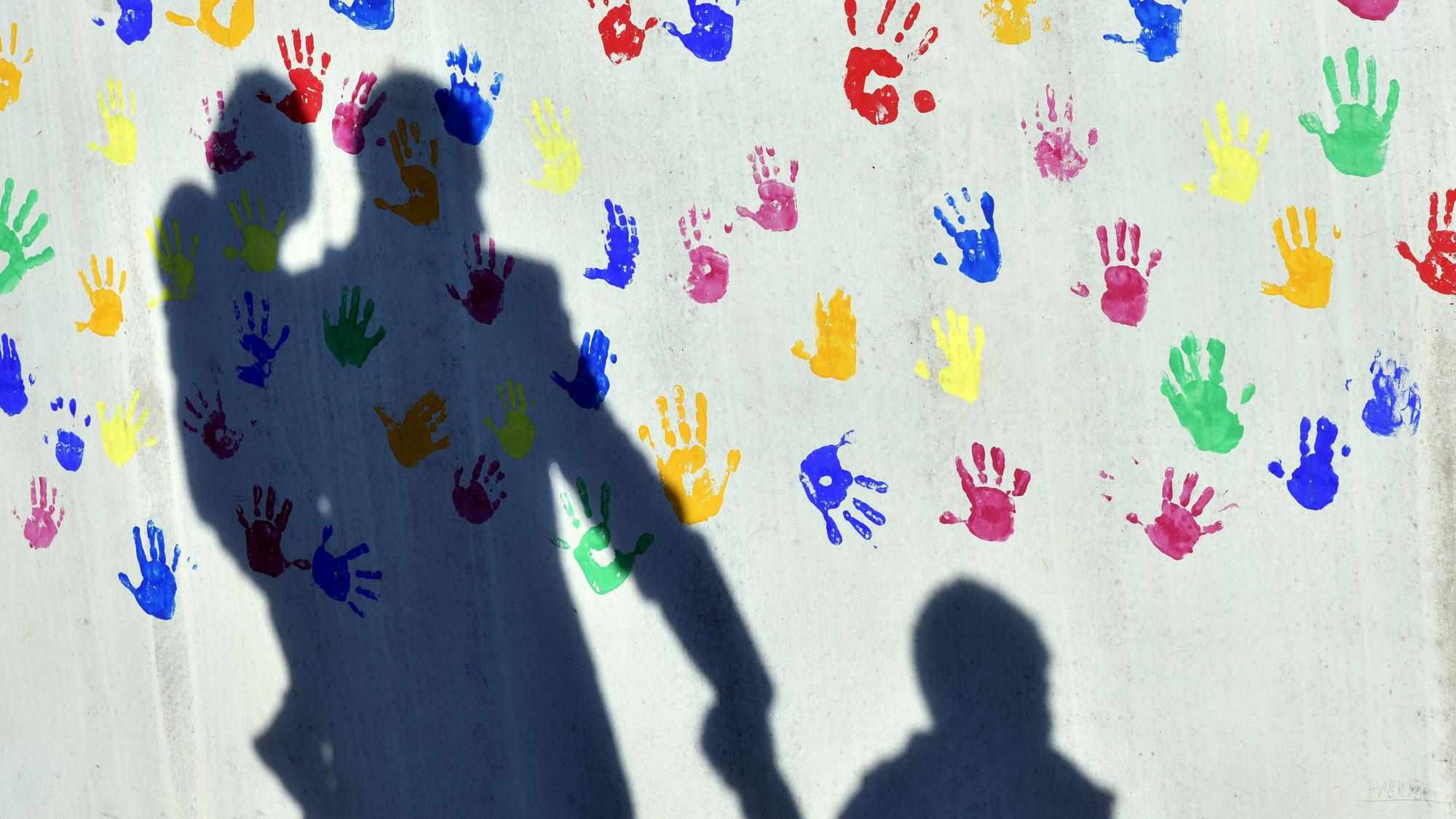 Ein Mann mit einem Kind auf dem Arm und einem an der Hand wirft einen Schatten auf eine mit bunten Handabdrücken bemalte Wand einer Kindertagesstätte.