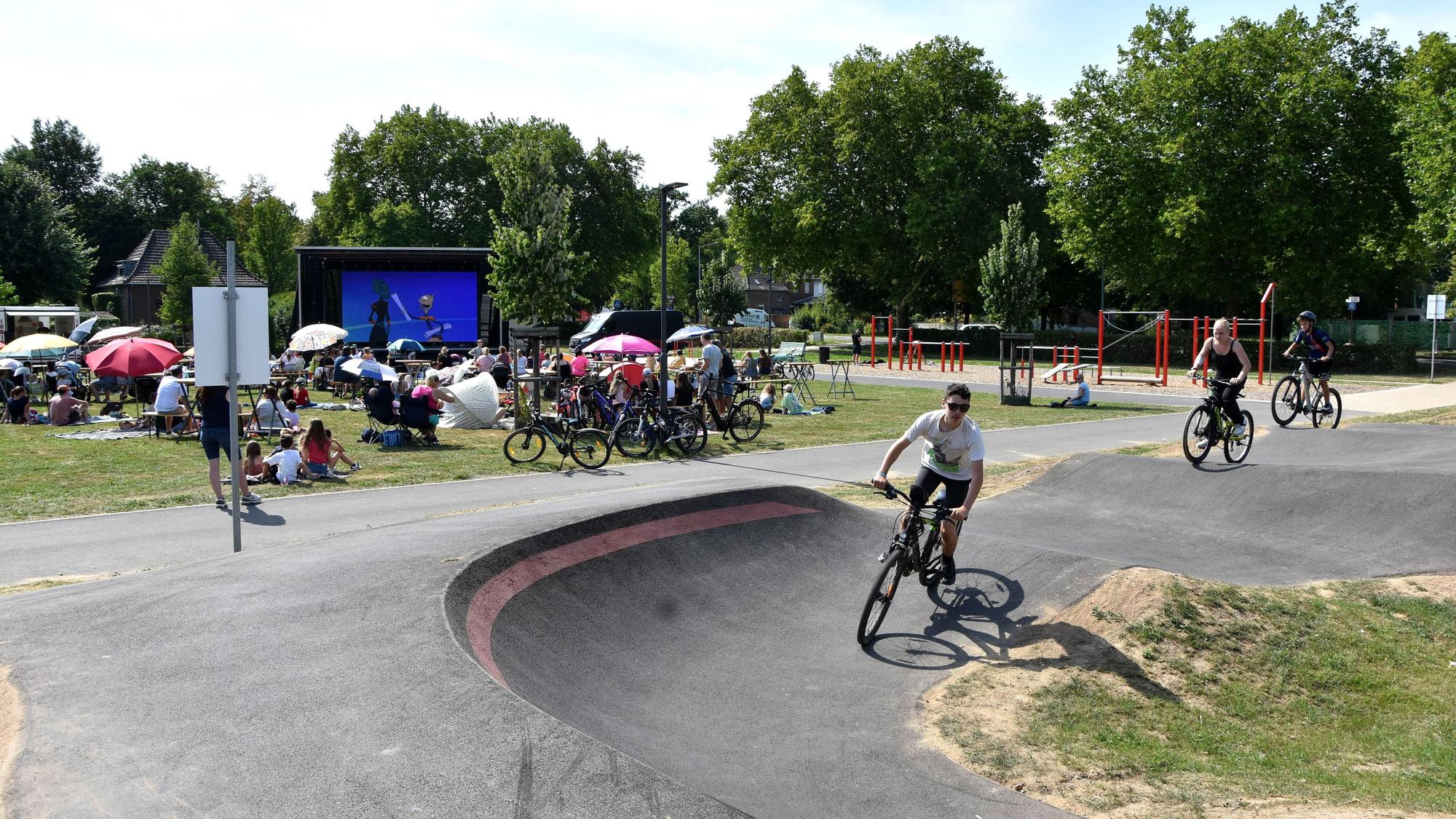 Jugendliche fahren auf ihren Mountainbikes auf der Strecke, im Hintergrund haben viele auf einer Wiese Platz genommen, um auf einer großen Leinwand den Film zu sehen