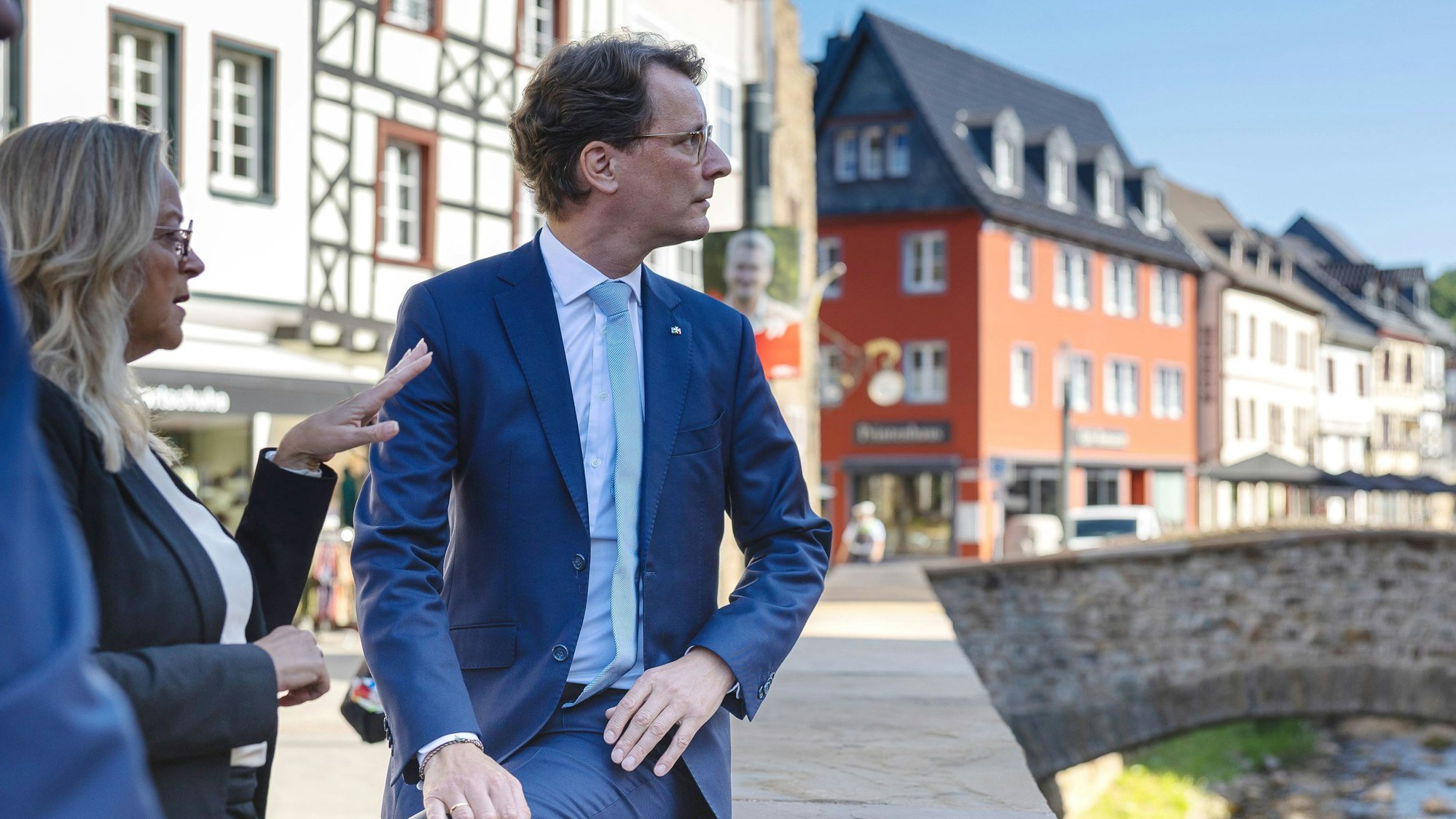 NRW-Ministerpräsident Hendrik Wüst sitzt in Bad Münstereifel auf einer Mauer an der Erft. Neben ihm steht Sabine Preiser-Marian und erklärt etwas.