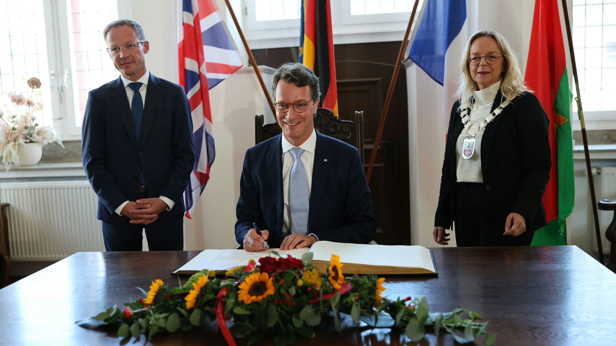 Ministerpräsident Hendrik Wüst schreibt ins Goldene Buch, neben ihm die Bürgermeisterin und der Staatssekretär. Im Hintergrund sind Fahnen zu sehen.