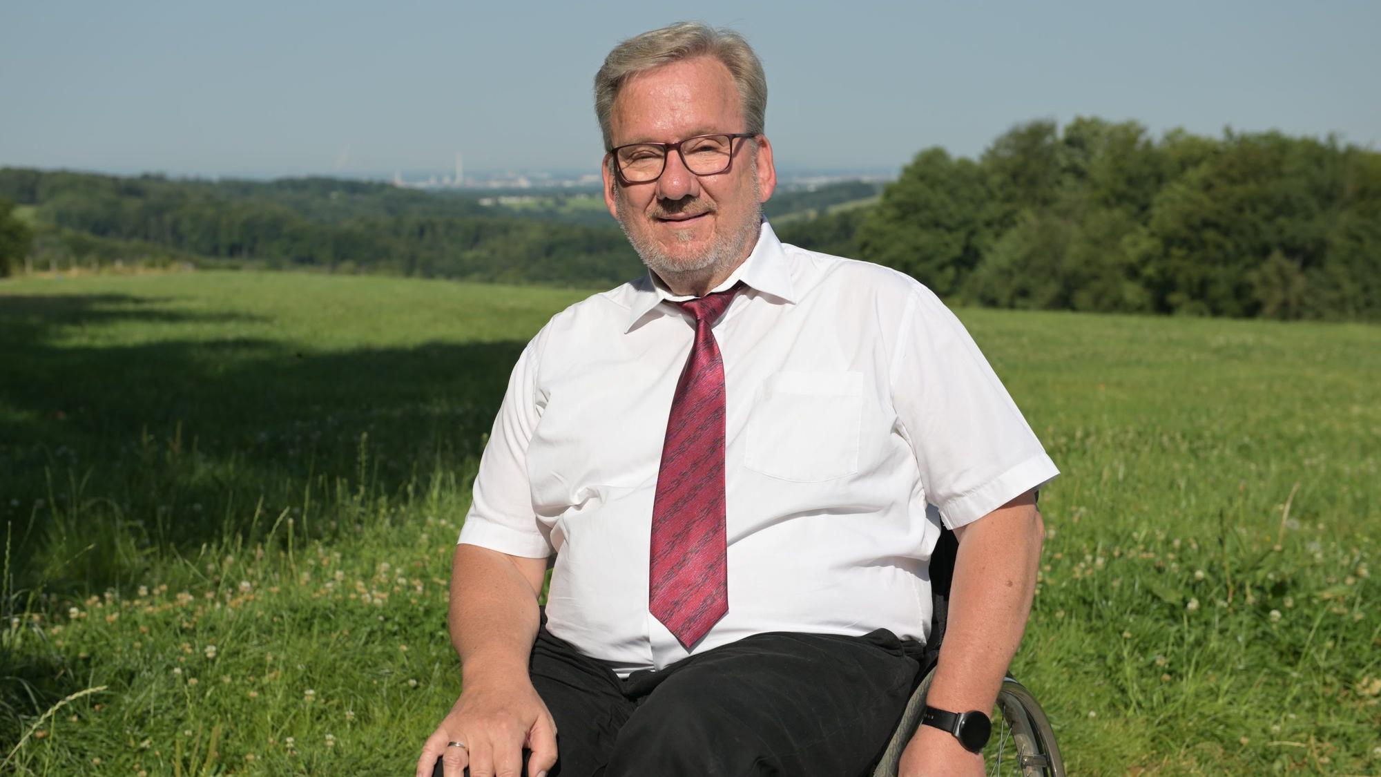 Ein Mann im Rollstuhl vor einer grünen Wiese, dahinter die Silhouette von Leverkusen.
