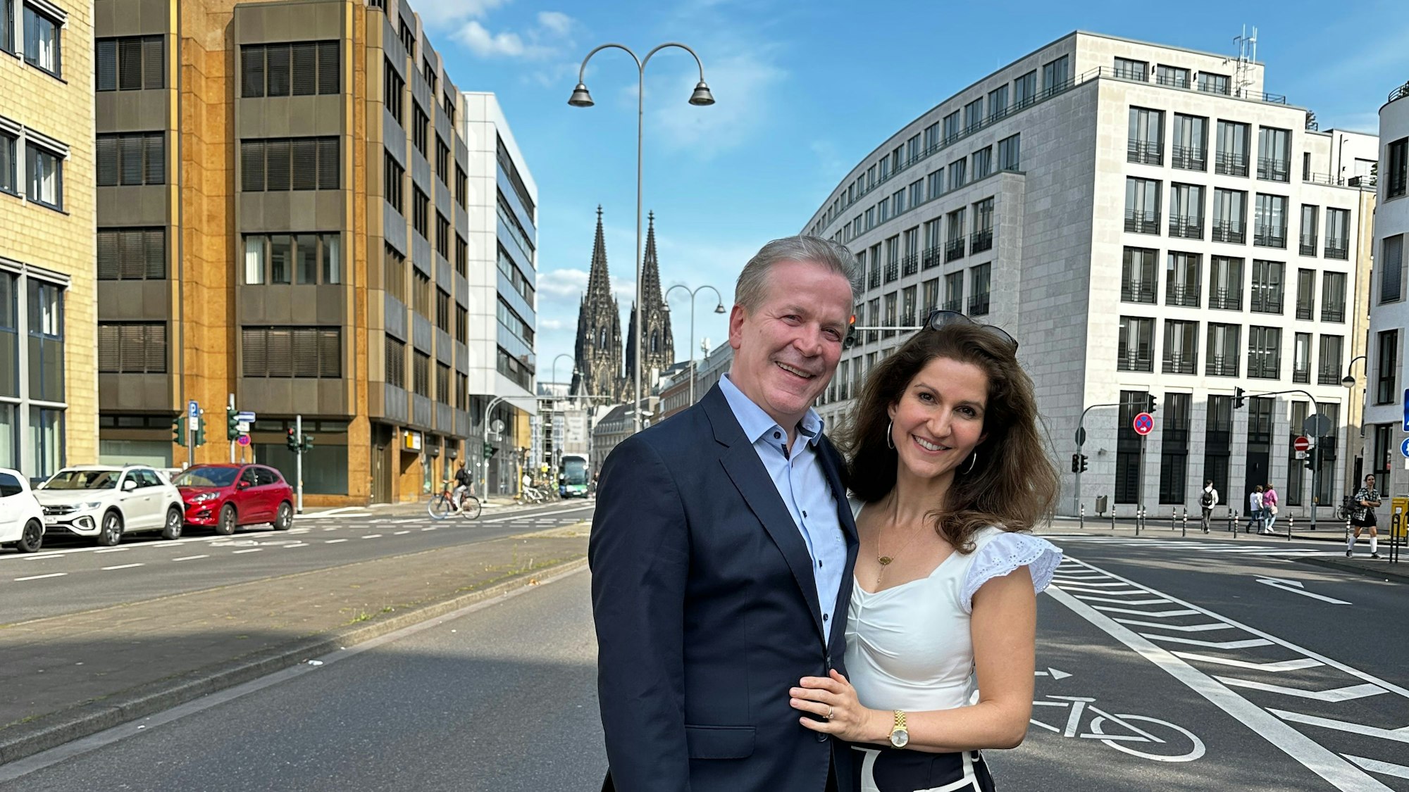 Auf der Gereonstraße stehen ein Mann und eine Frau und lächeln in die Kamera. Im Hintergrund ist der Dom zu sehen.