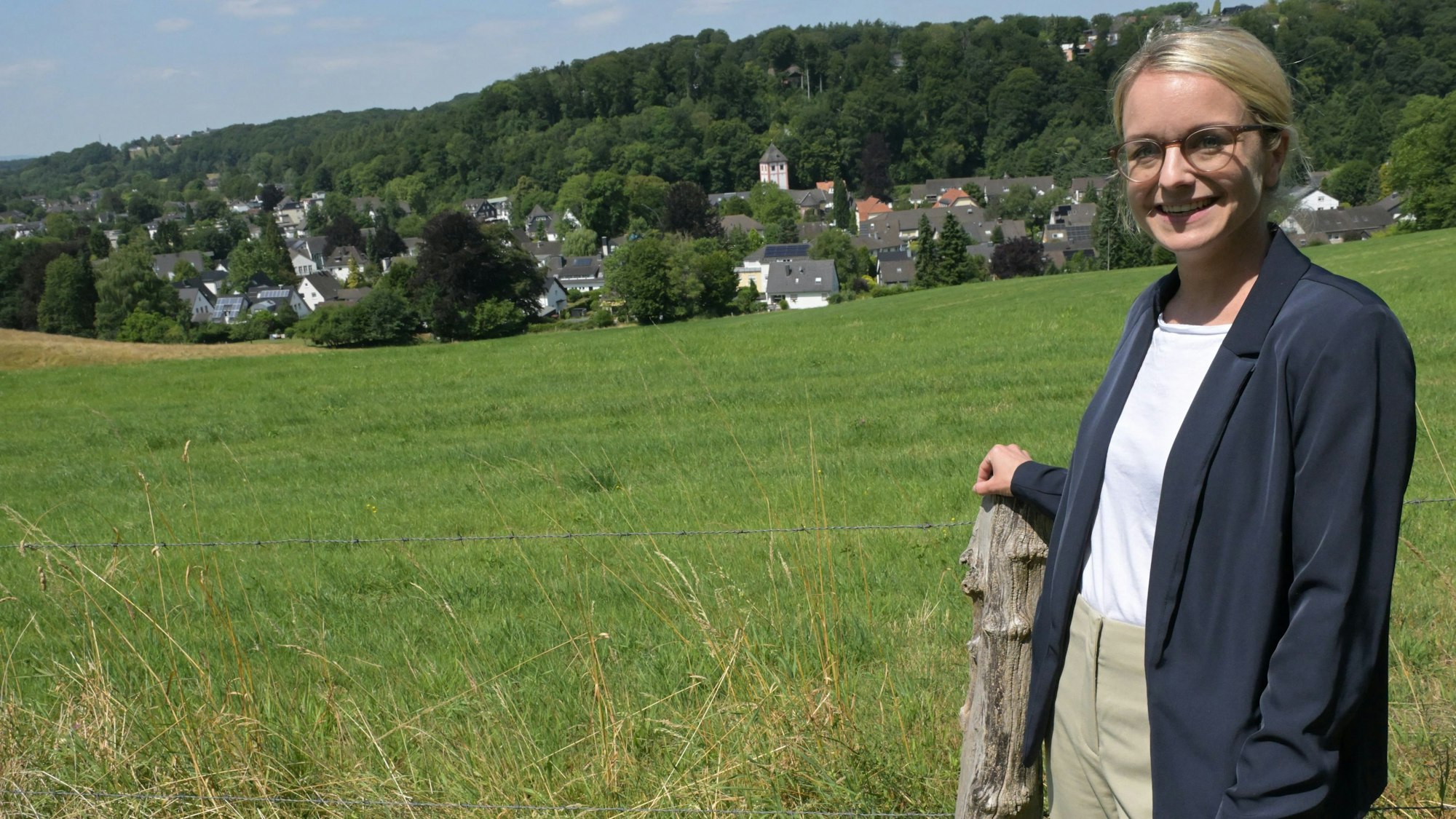 Eine junge Frau steht vor einer Wiese, unten im Tal ein kleines Örtchen mit Kirche.