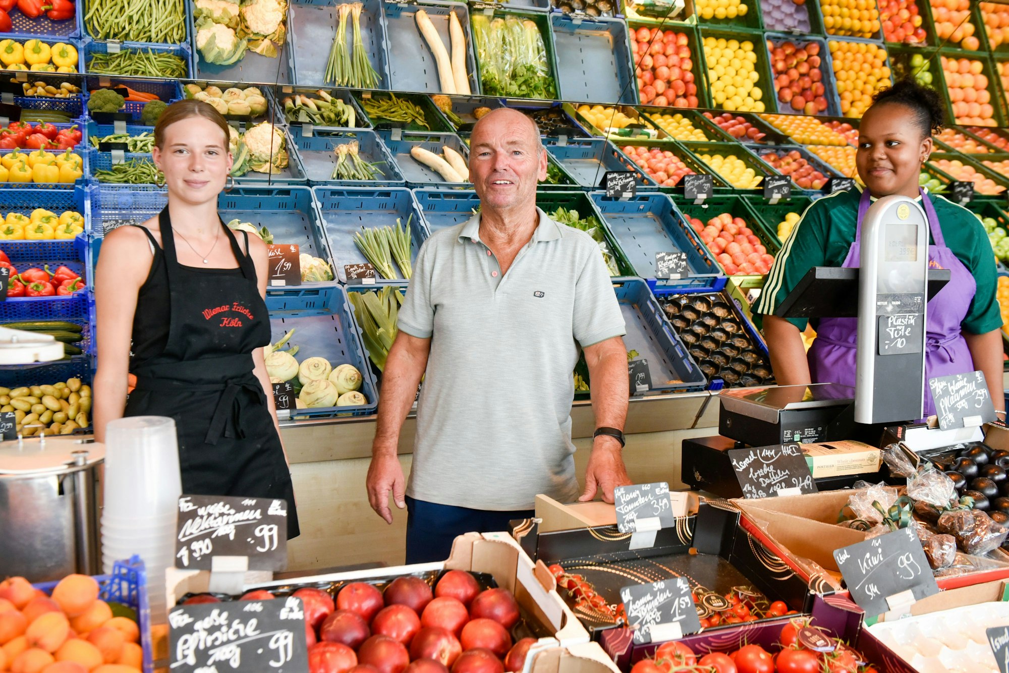 Auf dem Bild sind zwei Frauen und ein Mann vor einer reich gefüllten Obst- und Gemüsetheke zu sehen.