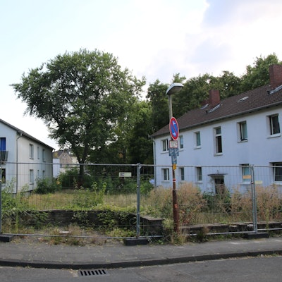 Drei zweigeschossige Gebäude mit einer Grünfläche, alles ist umgeben mit einem Bauzaun.