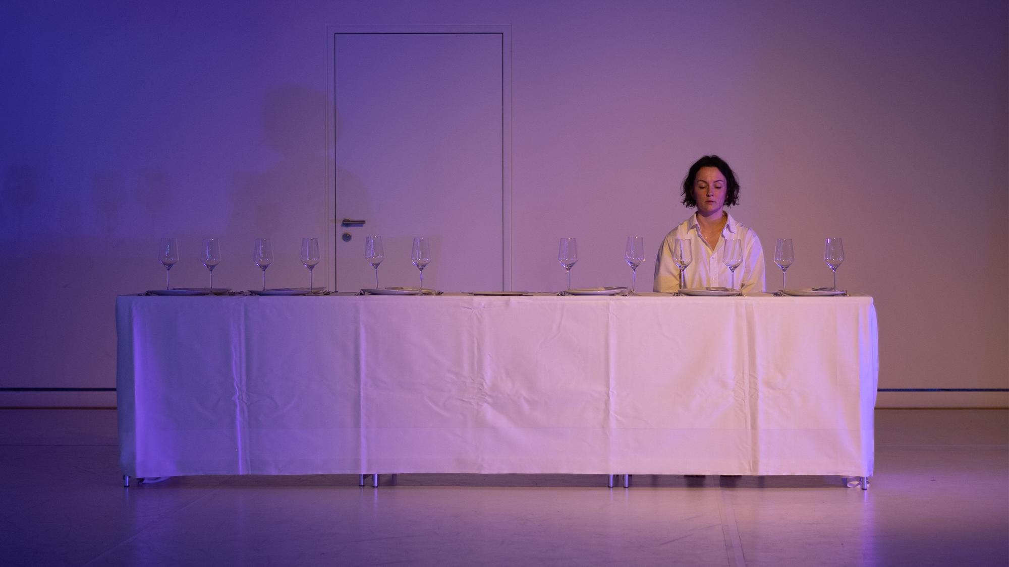 Eine Frau sitzt an einer langen, mit einer weißen Tischdecke gedeckten Tafel. Auf dem Tisch stehen 12 Weingläser.
