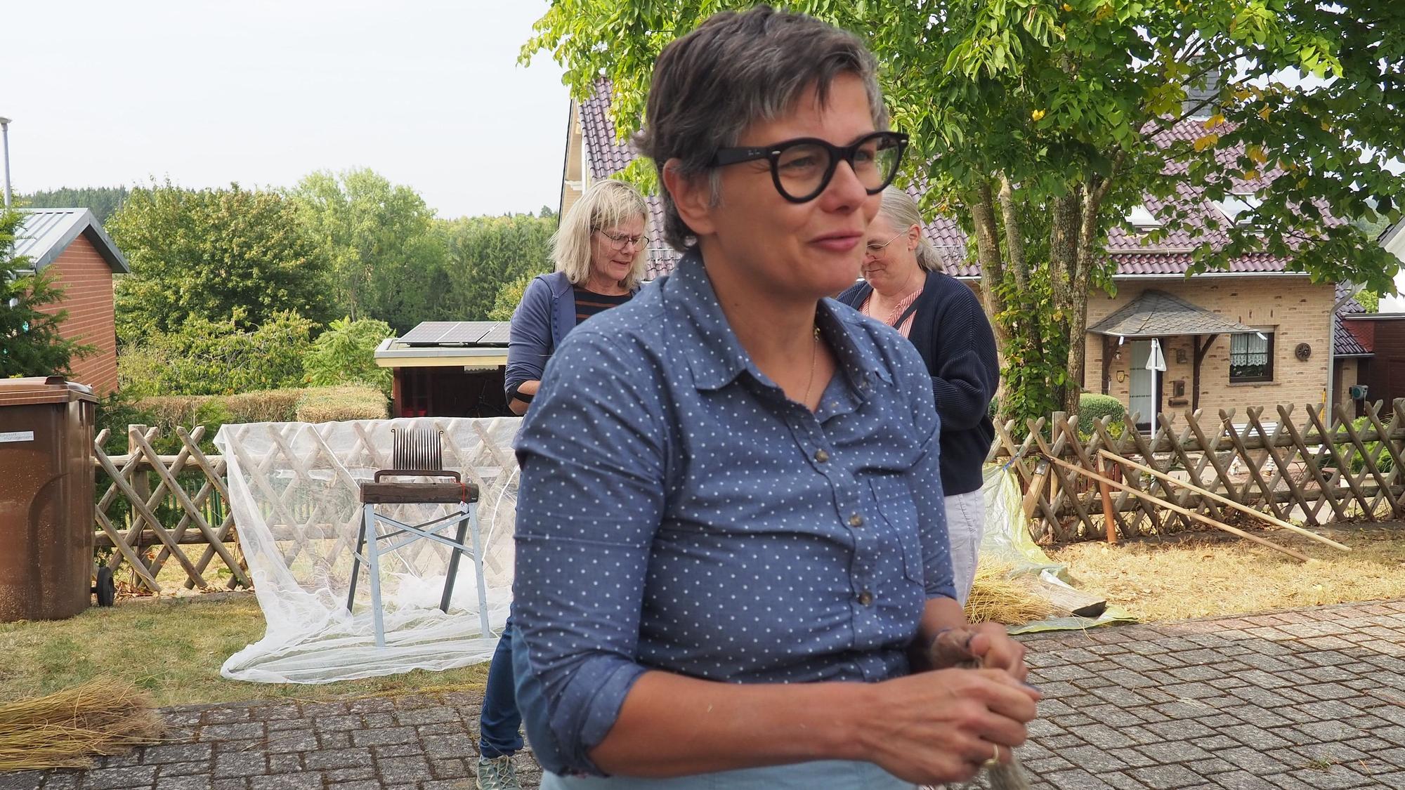 Christiane Seufferlein steht auf dem Hof vor dem Handwebmuseum in Rupperath. Im Hintergrund sind zwei Frauen zu sehen und die Vorrichtung, um Flachs zu rippeln.