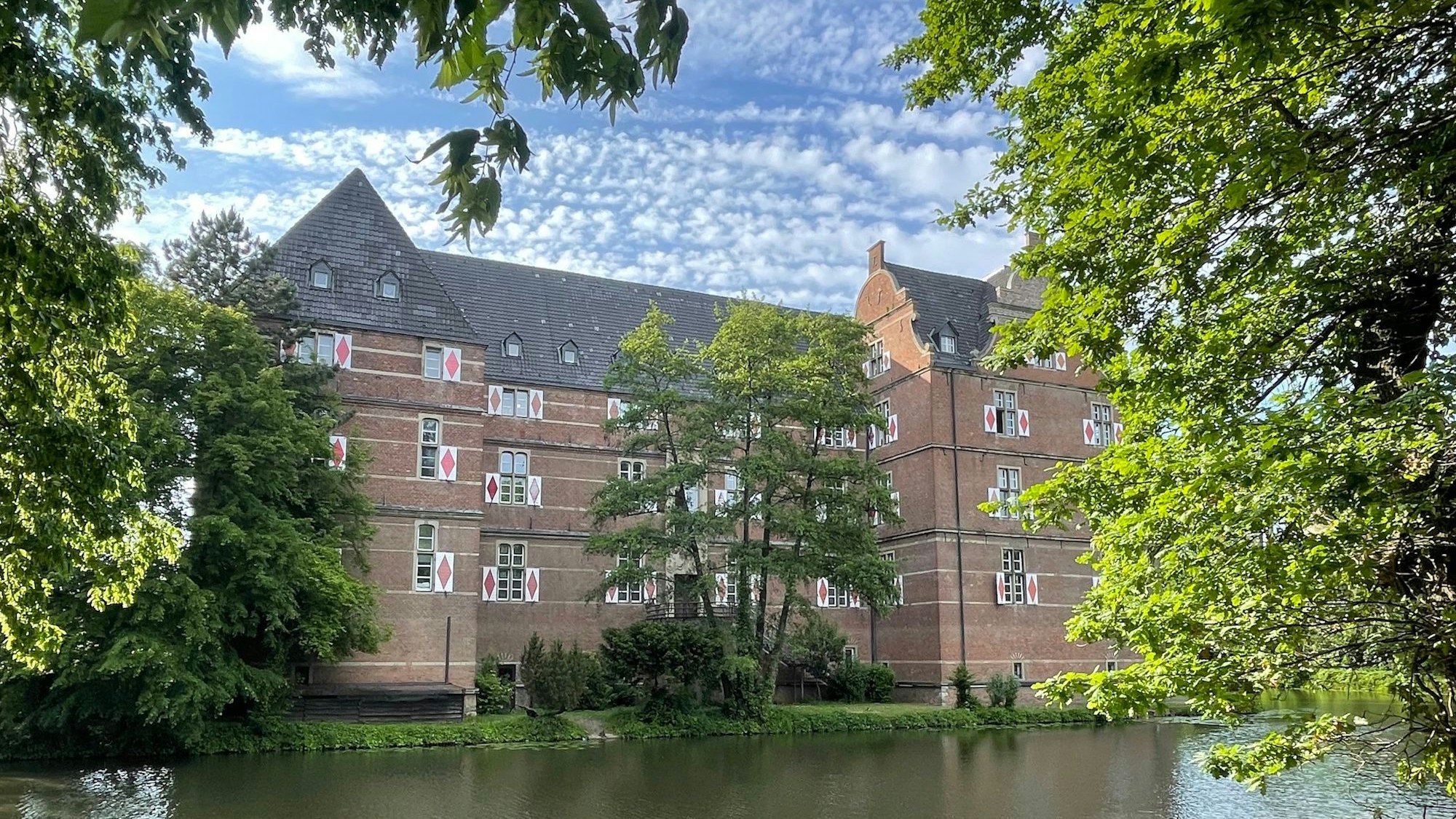 Zu sehen ist das Schloss Bedburg, es besteht aus rötlichem Stein. Vor dem Schloss ein Gewässer, umrahmt von Bäumen.