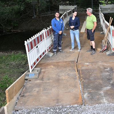 Zwei Männer und eine Frau stehen auf einer Behelfsbrücke aus Stahlplatten, die über einen Bach im Wald führt.