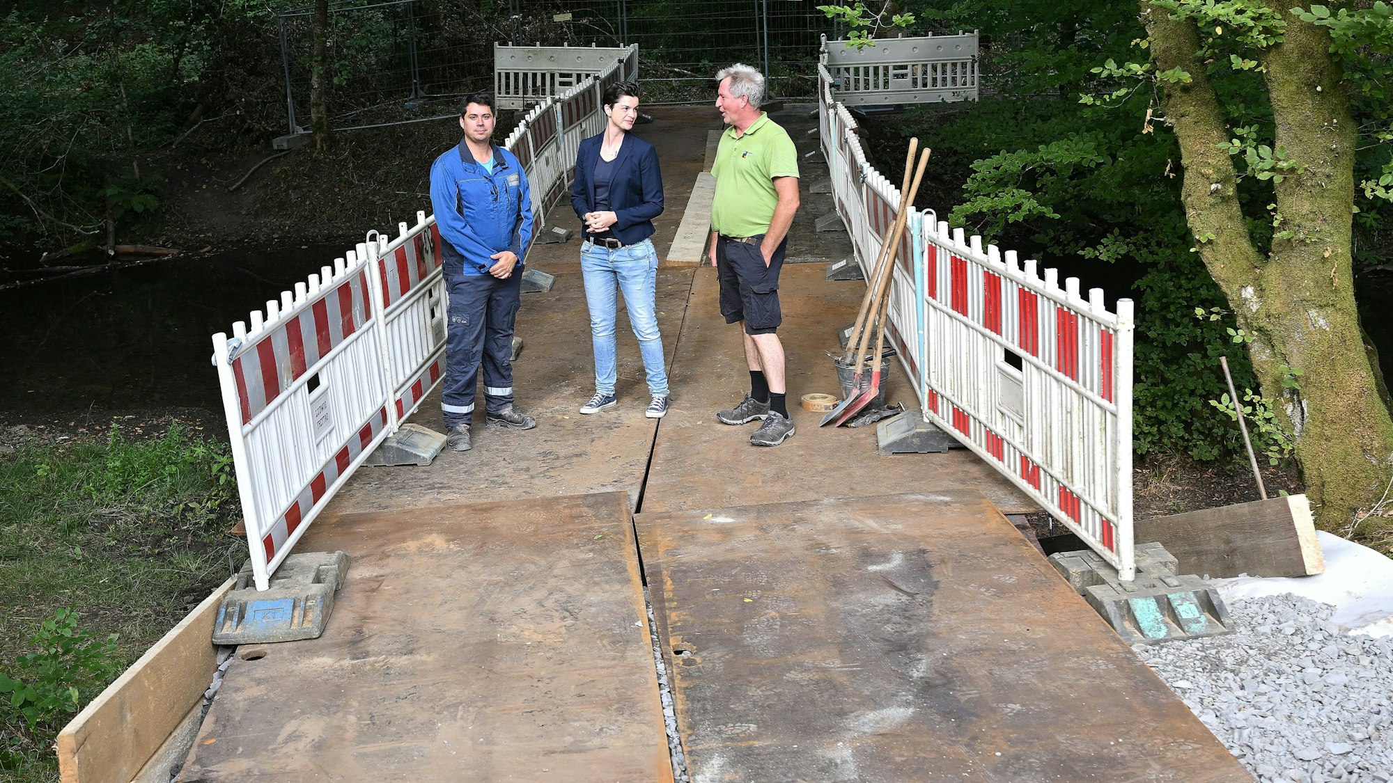 Zwei Männer und eine Frau stehen auf einer Behelfsbrücke aus Stahlplatten, die über einen Bach im Wald führt.