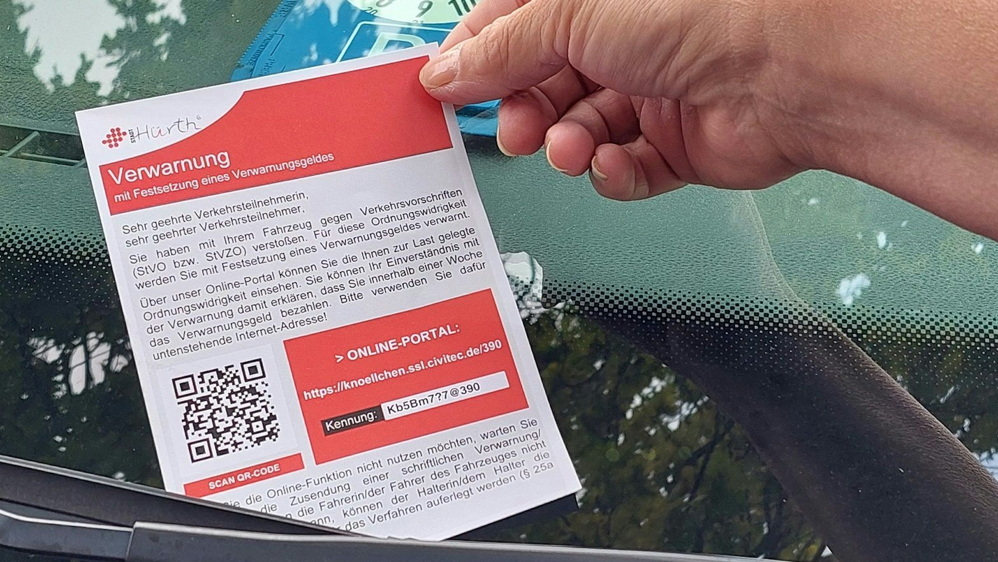 Eine Hand hält einen Verwarnungszettel, der hinter einem Scheibenwischer klemmt.