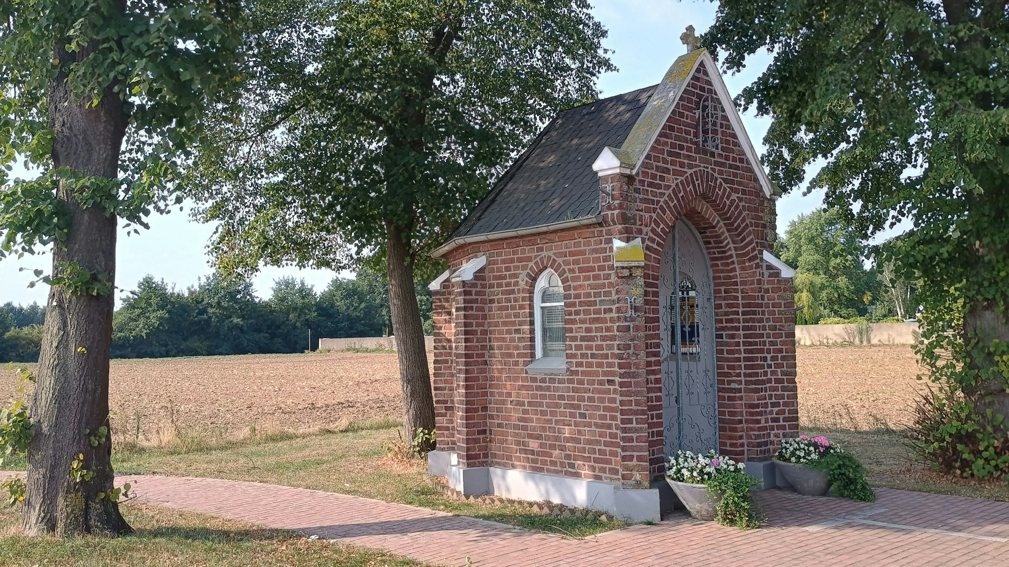 Die kleine Kapelle ist aus Backstein und hat ein spitzes Dach. Um sie herum stehen ein paar Bäume, vor ihr sind zwei Kübel mit Blumen.