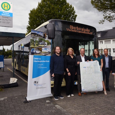 Fünf Menschen stehen am Bahnhof in Waldbröl vor einem Bus und halten ein Plakat hoch.
