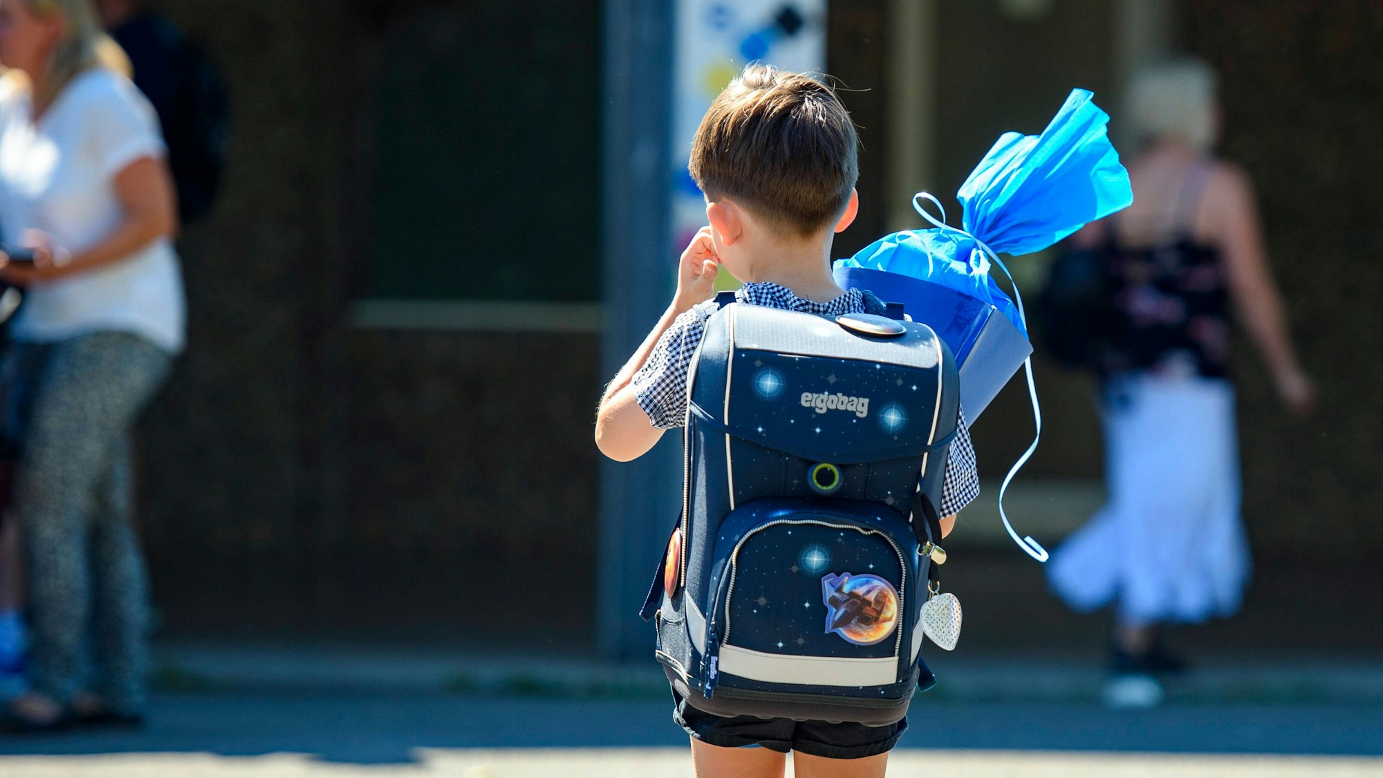 Ein Kind steht mit blauer Schultüte und Schultüte auf dem Schulhof