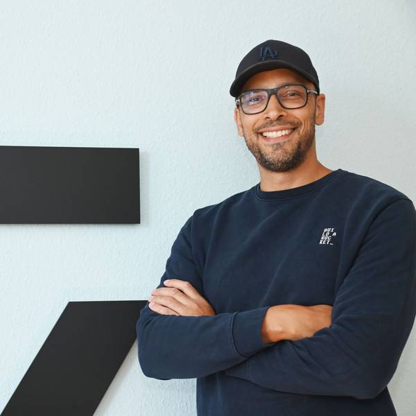 Alexander Albrecht, Brille, Cap, dunkelblauer Pullover, steht mit verschränkten Armen vor dem Logo seiner Brand und lächelt.