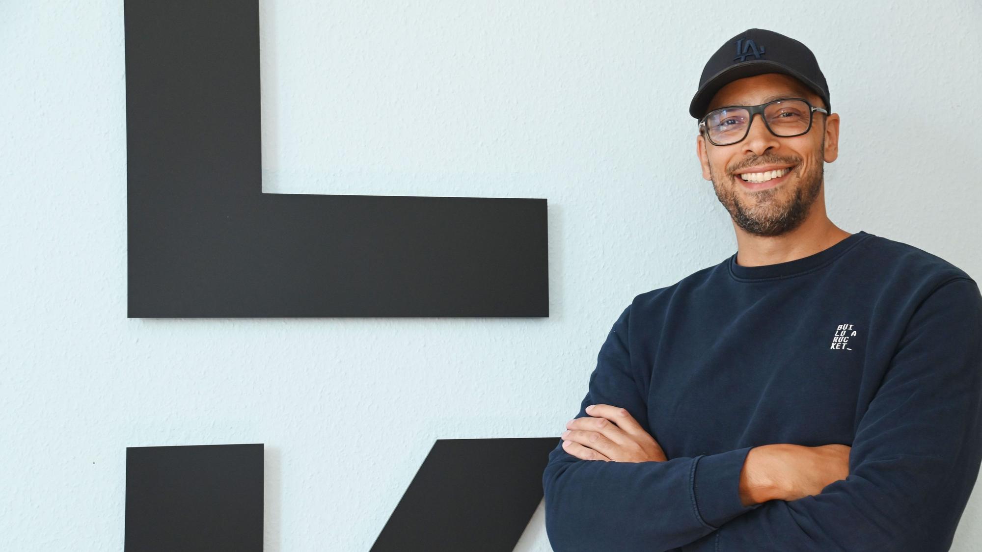 Alexander Albrecht, Brille, Cap, dunkelblauer Pullover, steht mit verschränkten Armen vor dem Logo seiner Brand und lächelt.