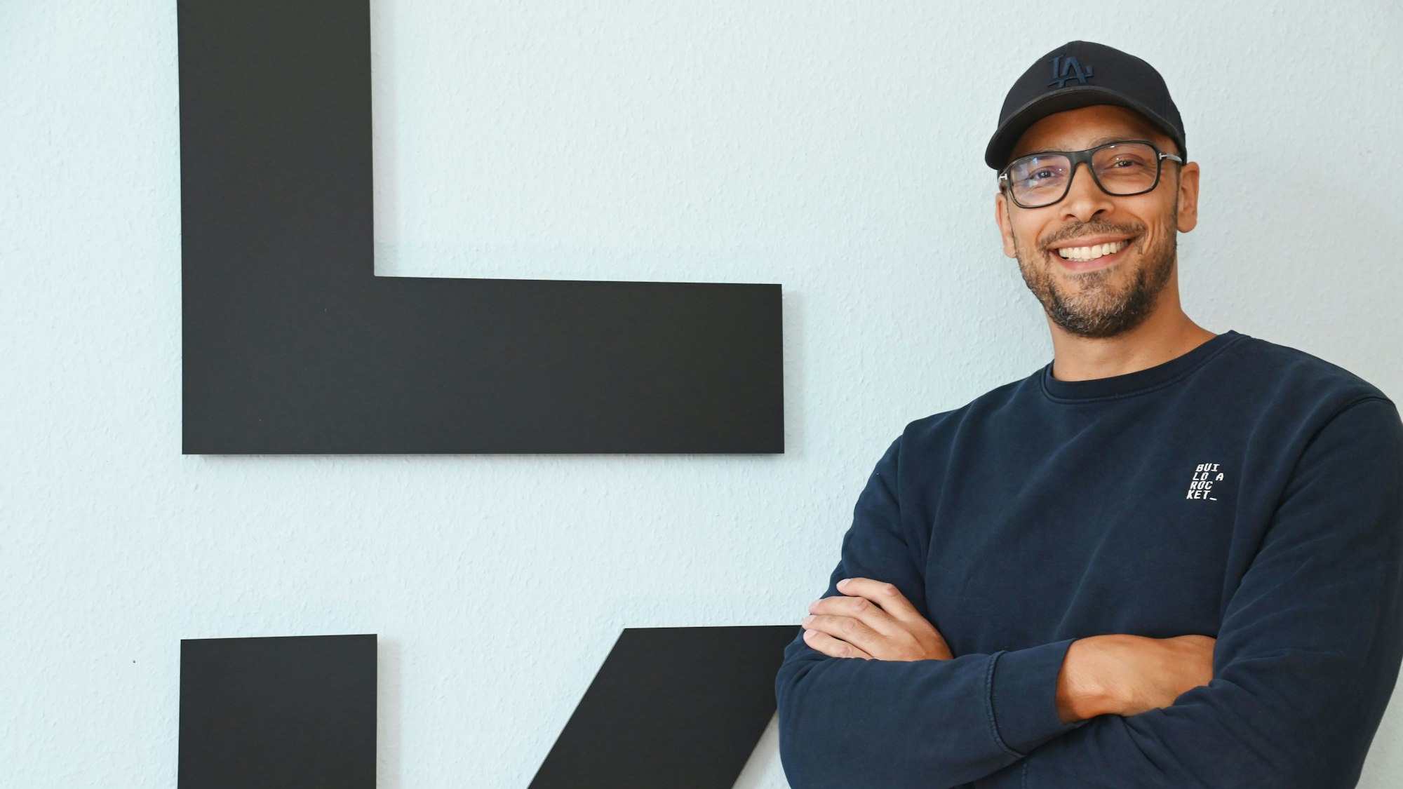 Alexander Albrecht, Brille, Cap, dunkelblauer Pullover, steht mit verschränkten Armen vor dem Logo seiner Brand und lächelt.