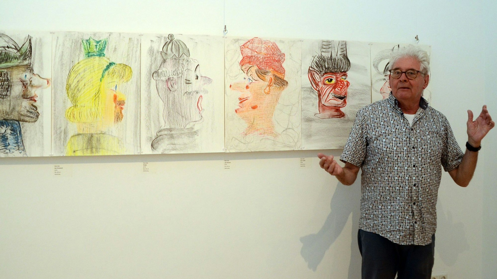 Ein Mann in einem Kurzarmhemd steht vor einer Reihe von bunten Zeichnungen, die an einer Wand hängen.