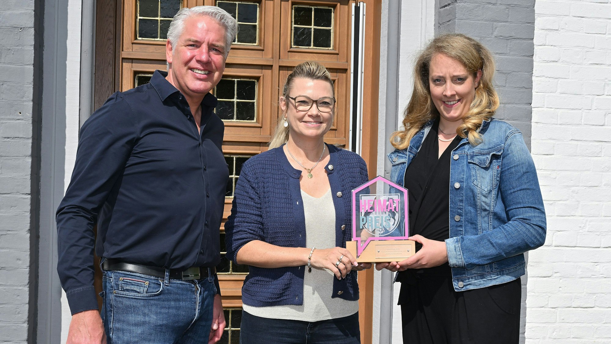 Ein Mann und zwei Frauen stehen vor der Tür des Odenthaler Rathauses und halten den Odenthaler Heimatpreis in der Hand.