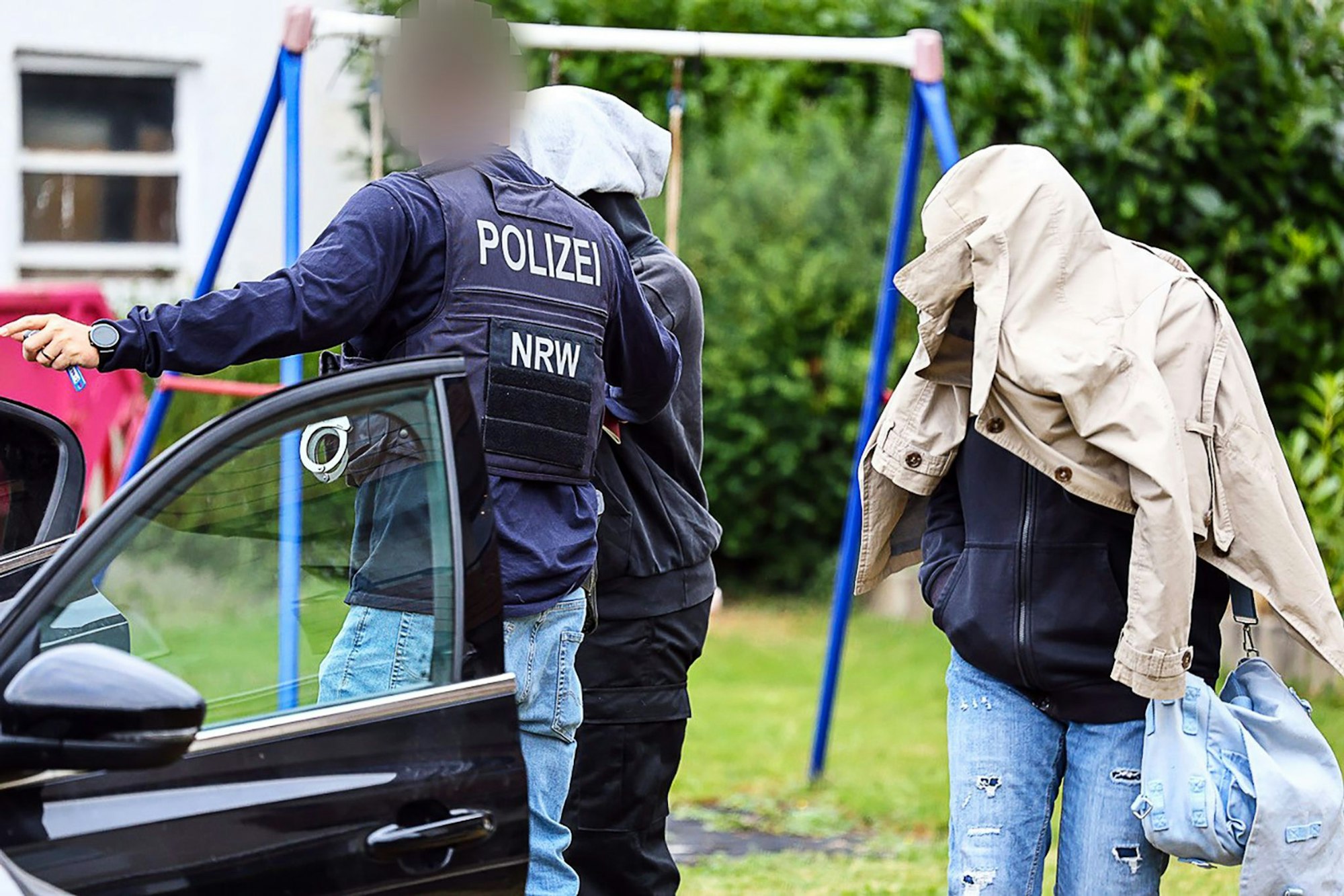 Ein Polizist führt zwei Personen in Herdecke, die ihre Gesichter verdecken, zu einem Auto.