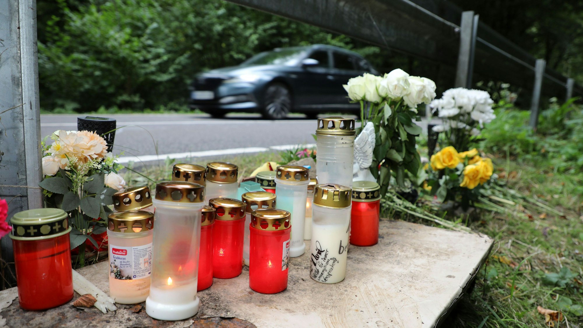 Kerzen und Blumen stehen unter einer Leitplanke der Straße, auf der am Samstag (16.8.) der Unfall bei Kürten-Dürscheid geschah.
