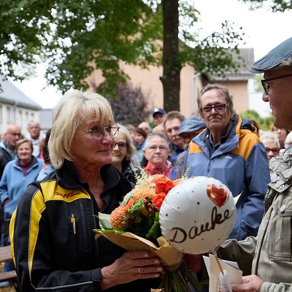 Christel Kastl steht links und hält einen Blumenstrauß und einen kleinen Ballon mit der Aufschrift „Danke“ in den Händen. Um sie herum stehen zahlreiche Menschen.