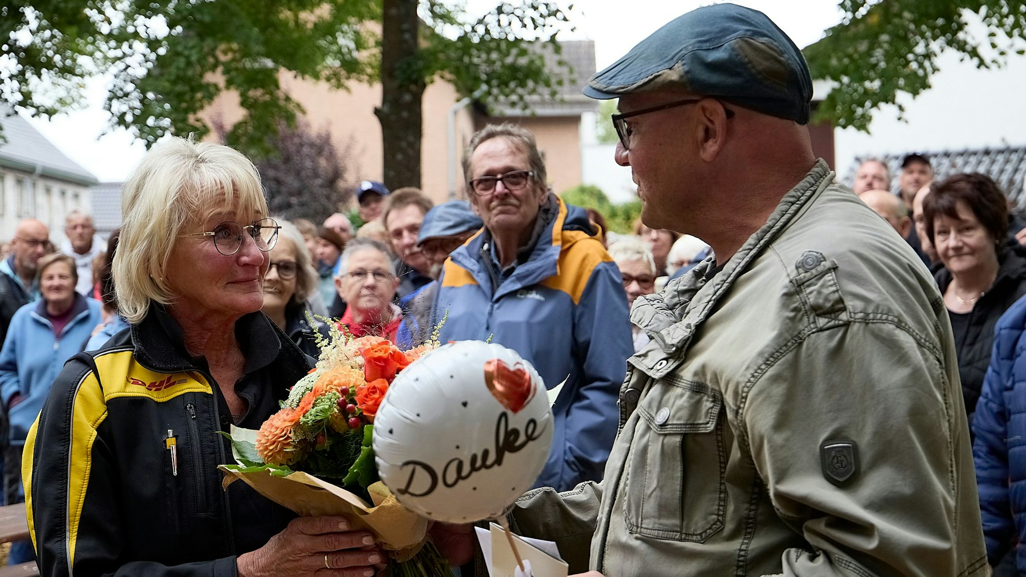 Christel Kastl steht links und hält einen Blumenstrauß und einen kleinen Ballon mit der Aufschrift „Danke“ in den Händen. Um sie herum stehen zahlreiche Menschen.