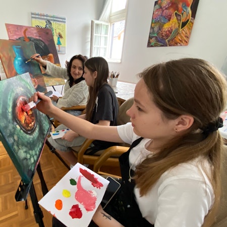 Olga Yankovska sitzt mit einem Mädchen an einer Staffelei, ein weiteres Mädchen an einer Staffelei sitzt daneben.