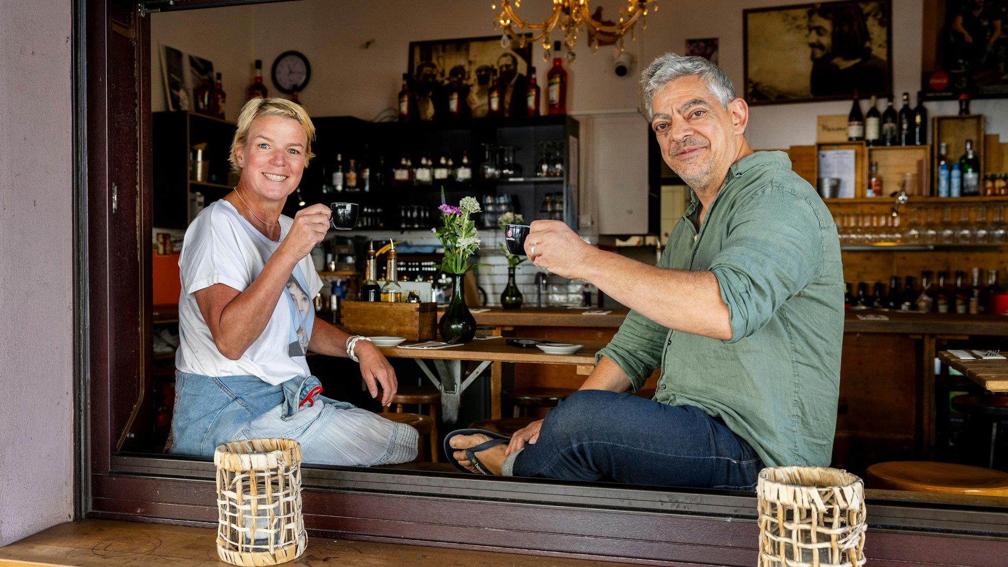Eine Frau mit blondem kurzem Haar sitzt einem Mann im grünen Hemd im offenen Fenster eines Restaurants gegenüber.