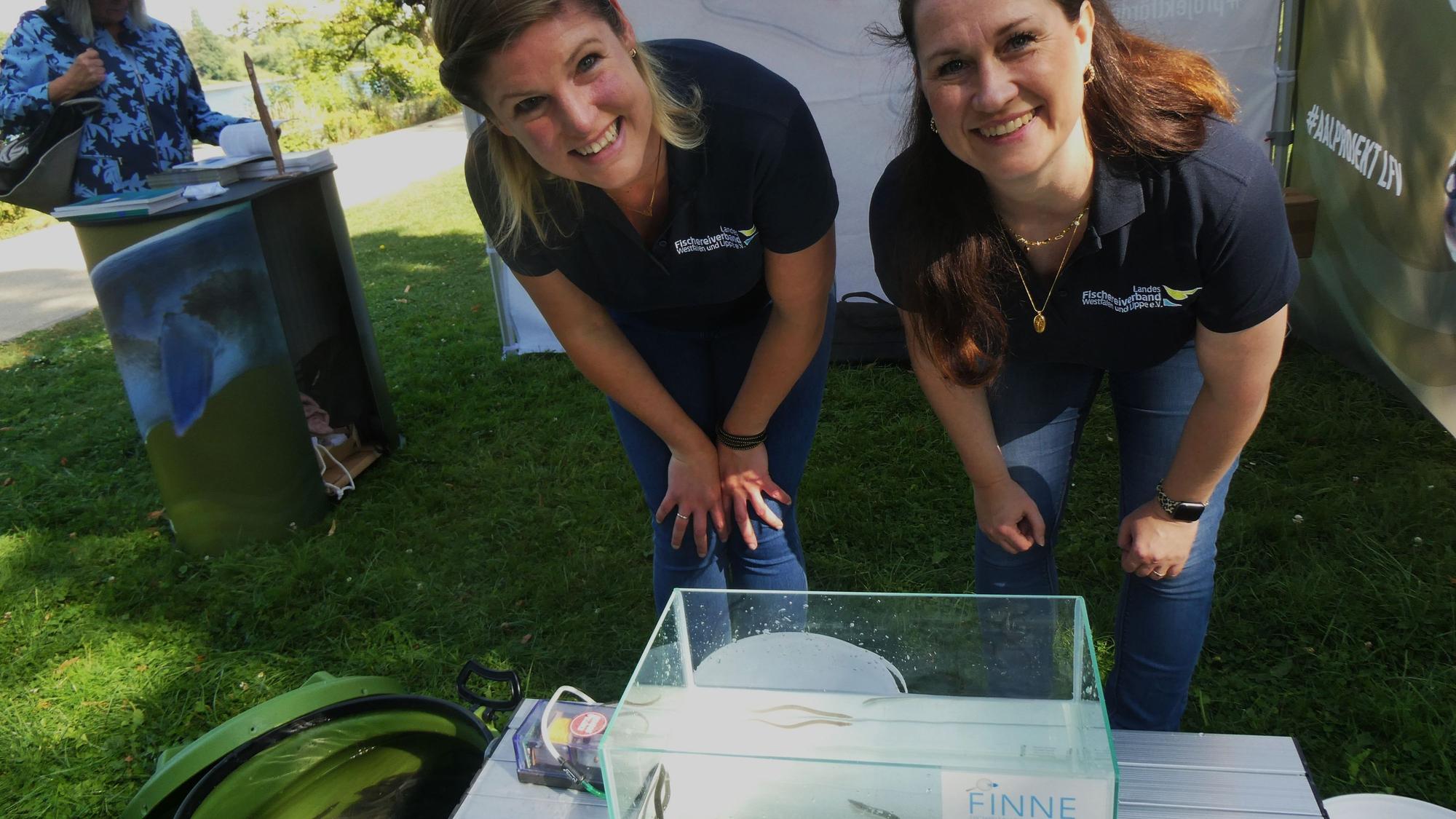Zwei Frauen mit einem kleinen Wasserbecken, in dem junge Aale schwimmen.