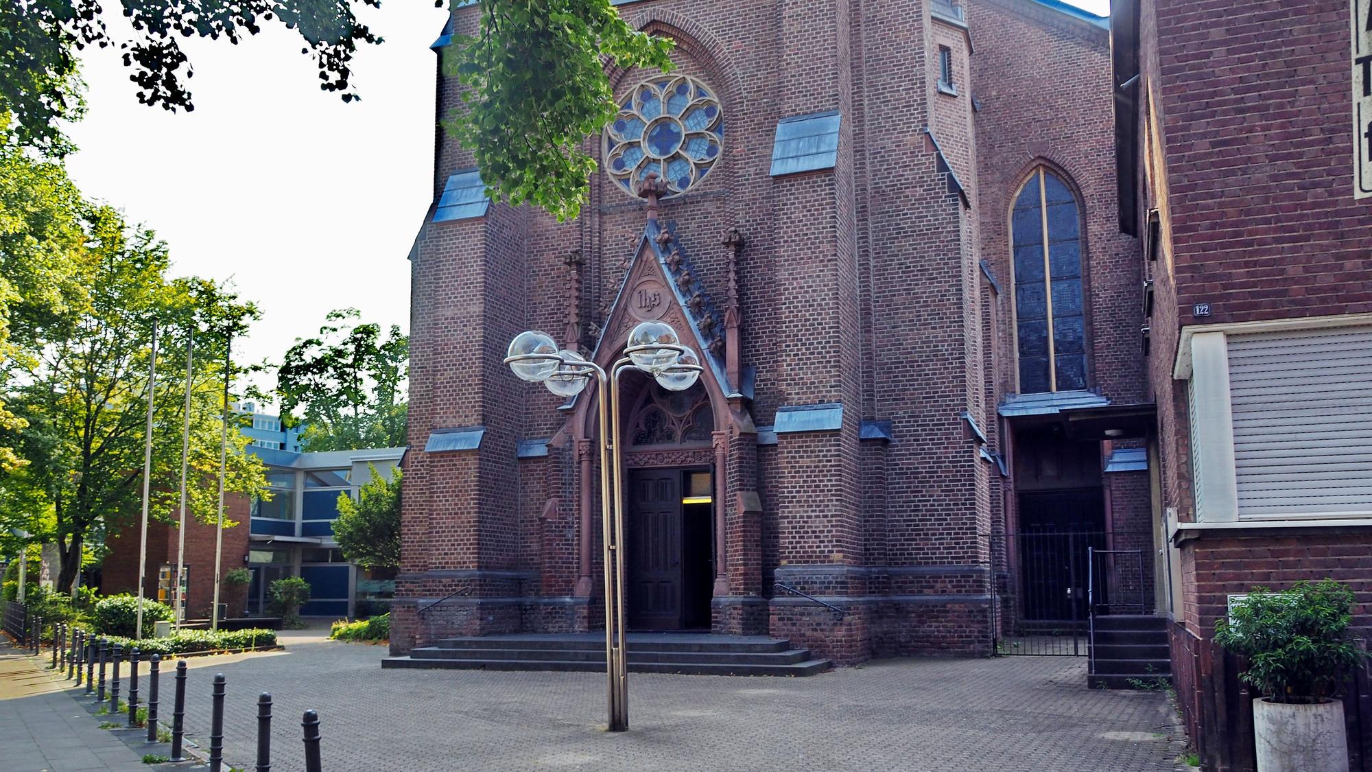 Vor einer Kirche steht eine Laterne, daneben ein Haus ebenso aus rötlichem Backstein gebaut.