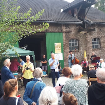 Ein Mann mit einem Mikro spricht vor zahlreichen Gästen. Im Hintergrund ist ein Hofgebäude zu sehen.