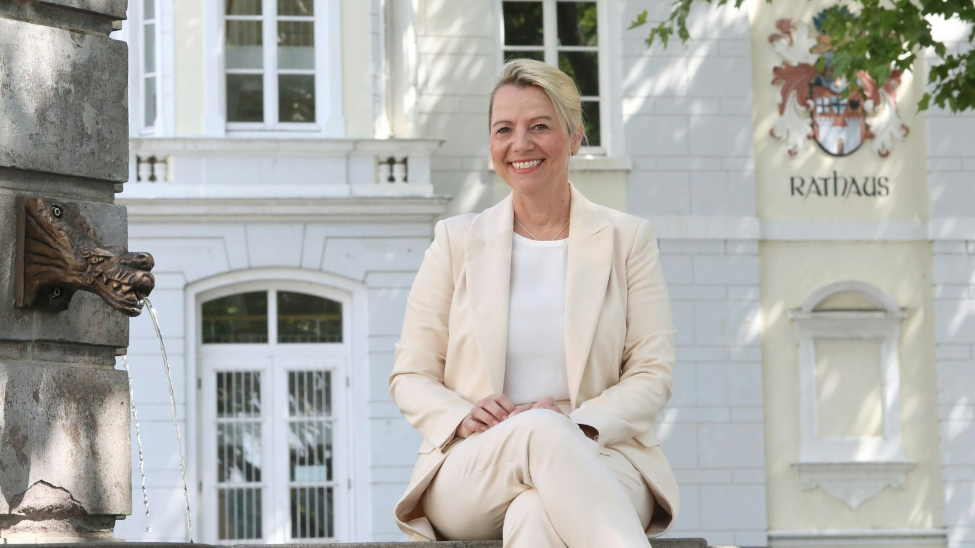 Eine Frau in beiger Kleidung sitzt an einem Brunnen, im Hintergrund auf einer Fassade der Schriftzug Rathaus sowie das Stadtwappen.