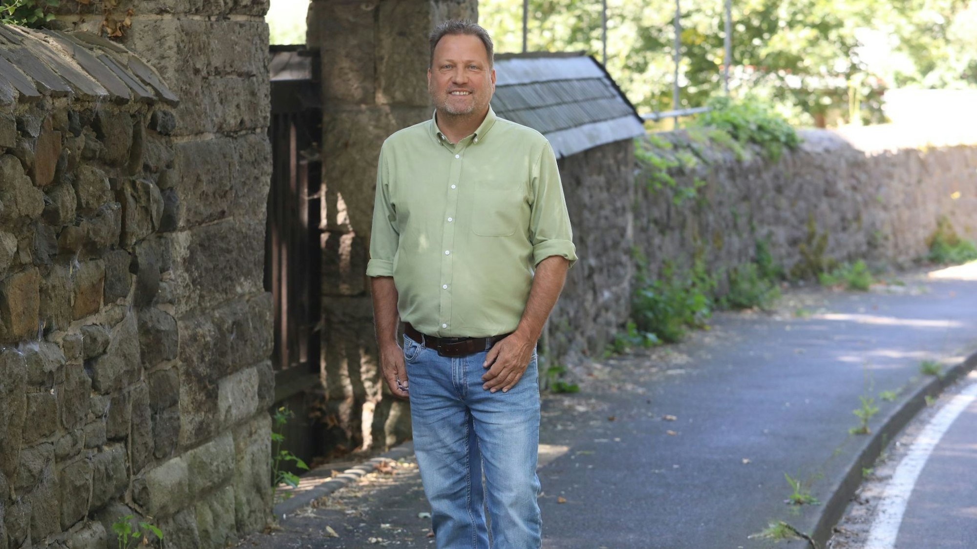 Ein Mann in Jeans und grünem Hemd auf einem Gehweg neben einer Bruchsteinmauer.