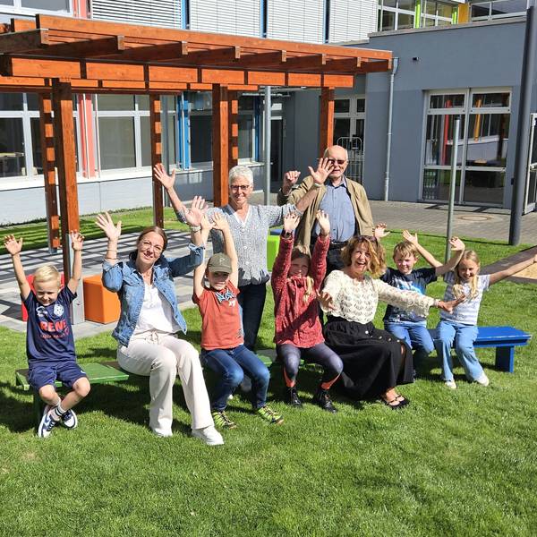 Kinder und Erwachsene sitzen auf Bänken vor der Pergola und winken mit den Händen.