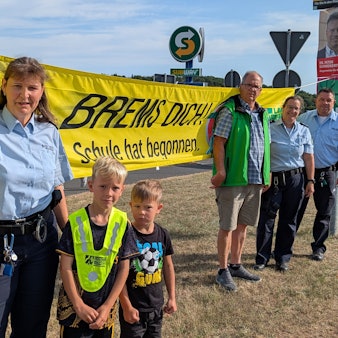 Vier Erwachsene und zwei Kinder stehen von einem Banner mit der Aufschrift „Brems Dich! Schule hat begonnen“.