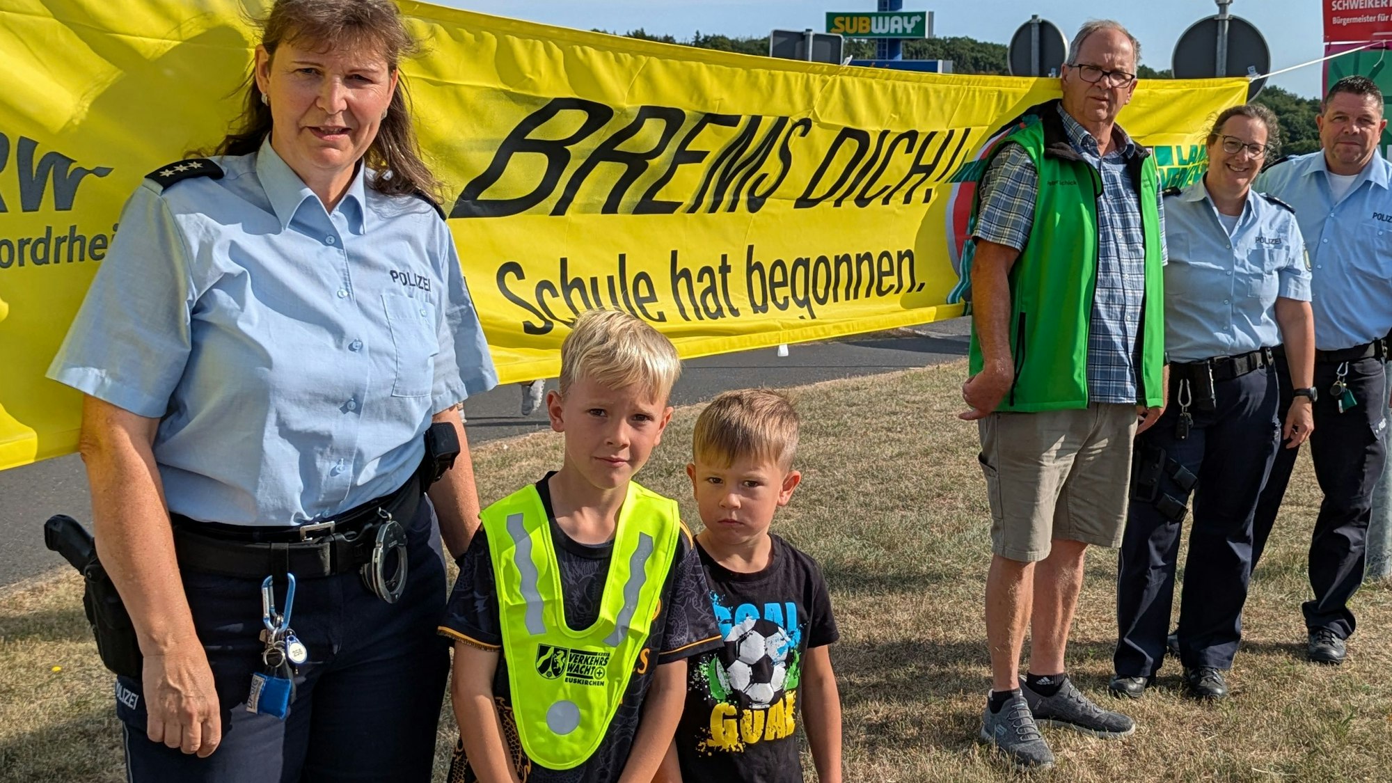 Vier Erwachsene und zwei Kinder stehen von einem Banner mit der Aufschrift „Brems Dich! Schule hat begonnen“.