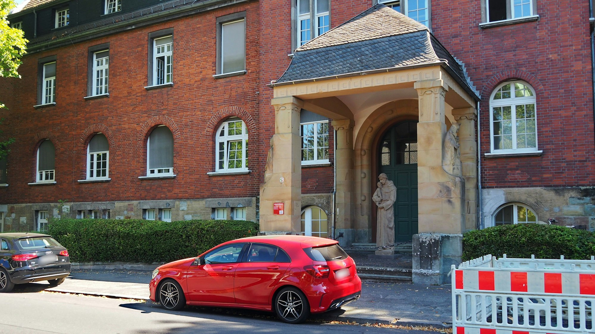 Vor einem großen Backsteingebäude mit stattlichem Säulen-Portal parken Autos auf dem Bürgersteig.