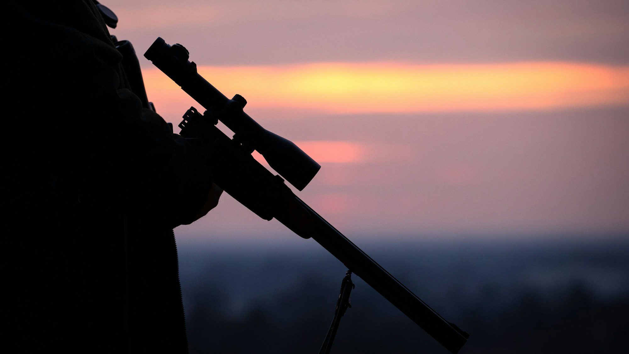 Ein Mann hält ein Gewehr mit einem Zielfernrohr vor einem Abendhimmel in der Hand.