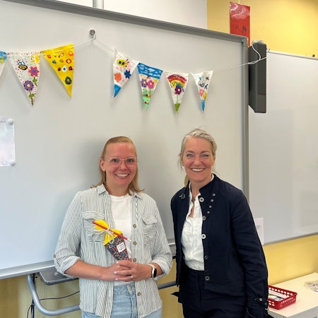 Die beiden stehen nebeneinander vor einem Whiteboard in einem Köassenzimmer.