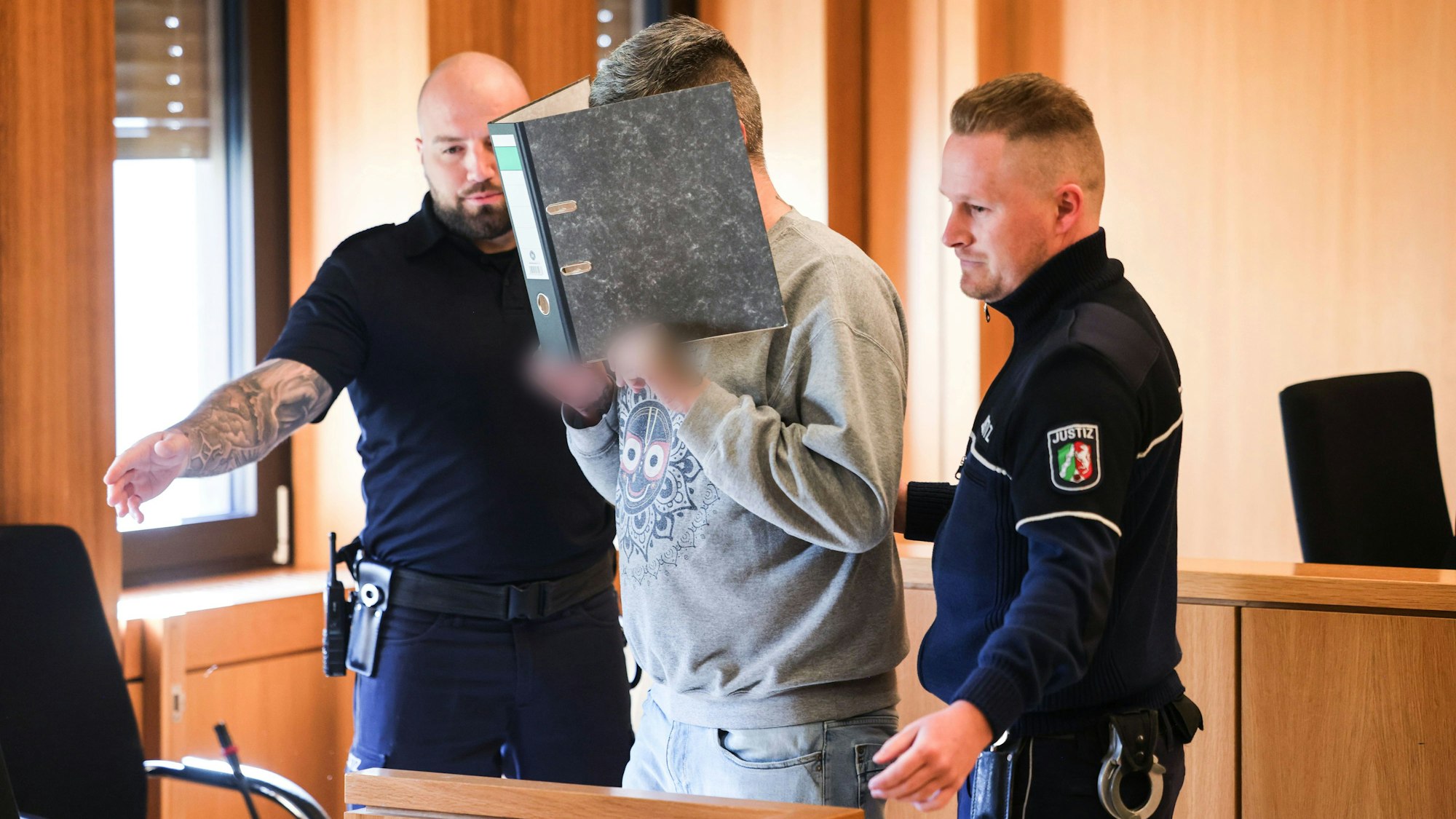 ARCHIV - 24.03.2025, Nordrhein-Westfalen, Aachen: Der angeklagte Krankenpfleger hält sich einen Aktenordner vor sein Gesicht, während er von Justizangestellten zu seinem Platz gebracht wird. Im Mordprozess gegen einen Krankenpfleger vor dem Landgericht Aachen haben ehemalige Kolleginnen von abfälligen Bemerkungen des Angeklagten über Patienten berichtet. (zu dpa: «Pfleger wegen Mordes in neun Fällen vor Gericht») Foto: Oliver Berg/dpa +++ dpa-Bildfunk +++