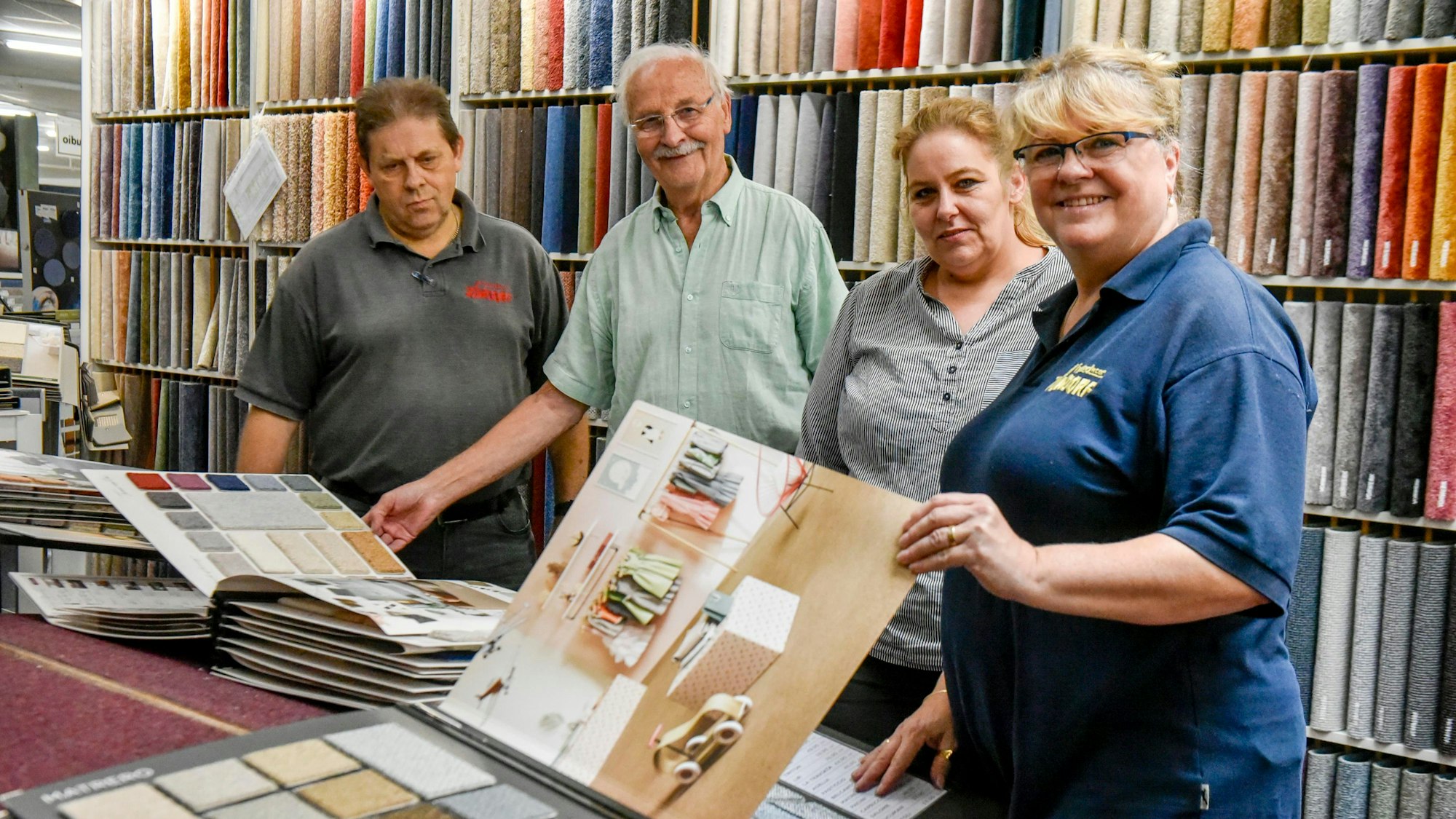 Auf dem Bild sind zwei Männer und zwei Frauen vor einem Regal mit Stoffmustern zu sehen, die Bücher mit Stoffproben präsentieren.