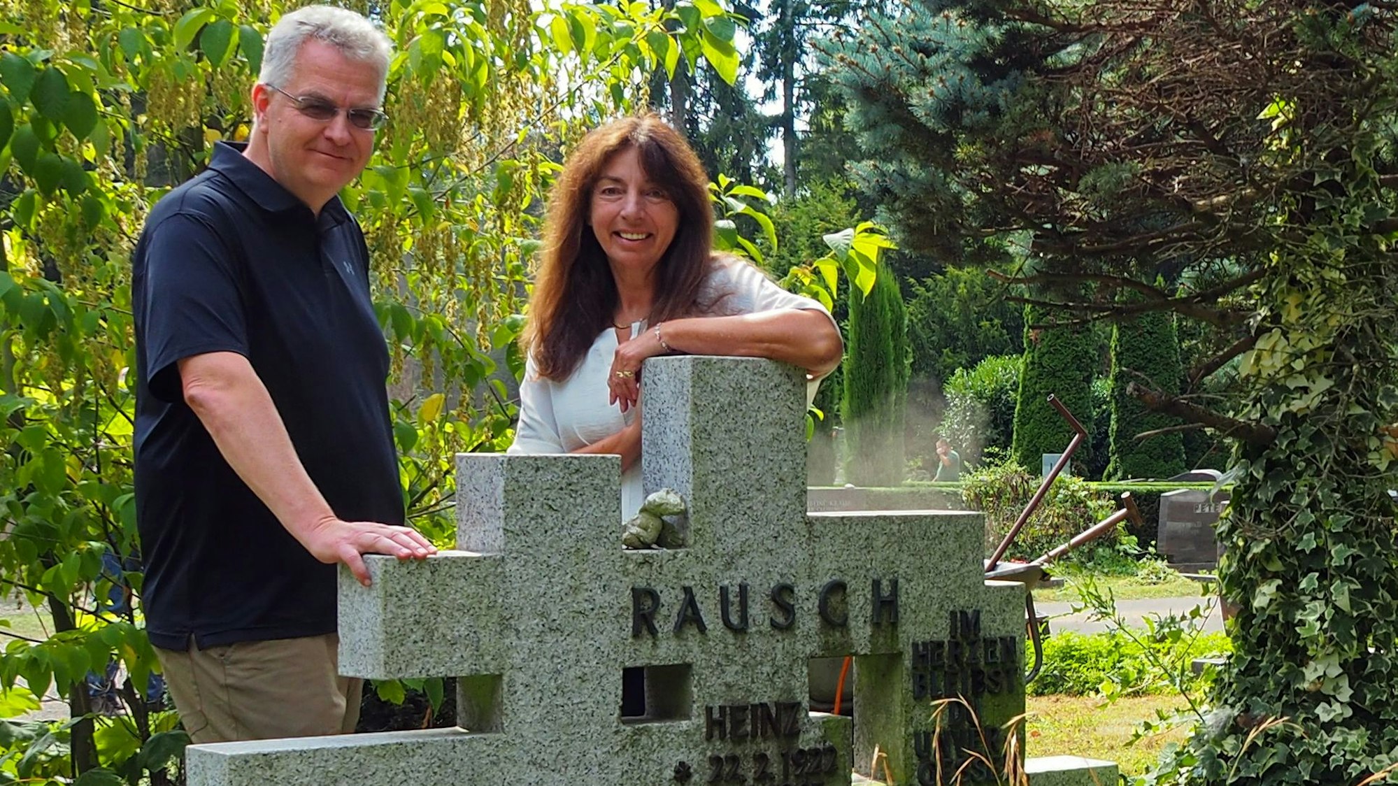 Ein Mann und eine Frau stehen neben einem grauen Grabstein auf einem Friedhof.
