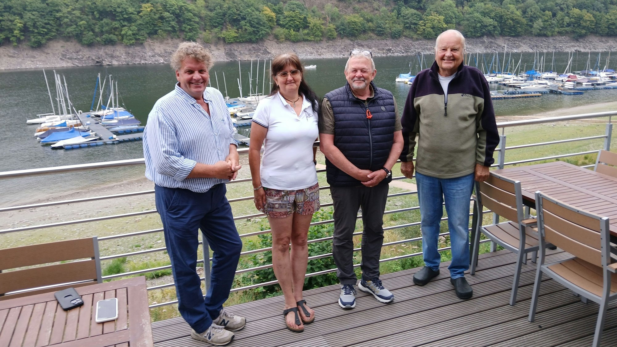 Drei Männer und eine Frau stehen auf dem Balkon des Vereinsheims, hinter ihnen der See.